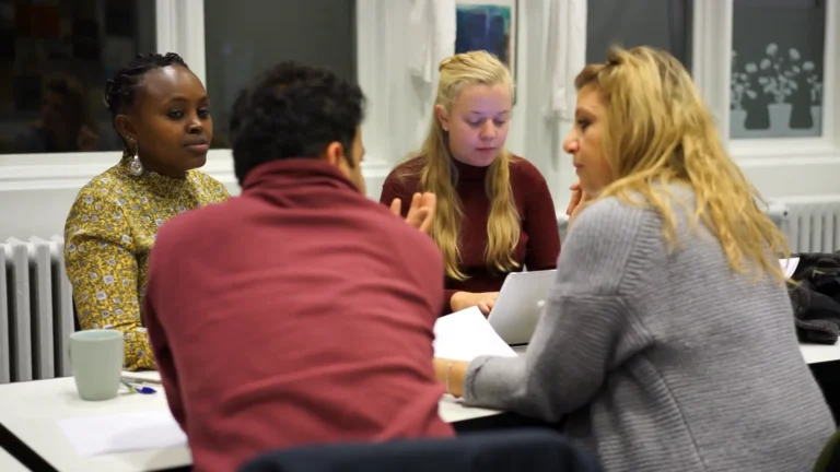 Fire personer diskuterer ved et bord og er i en alvorlig stemning